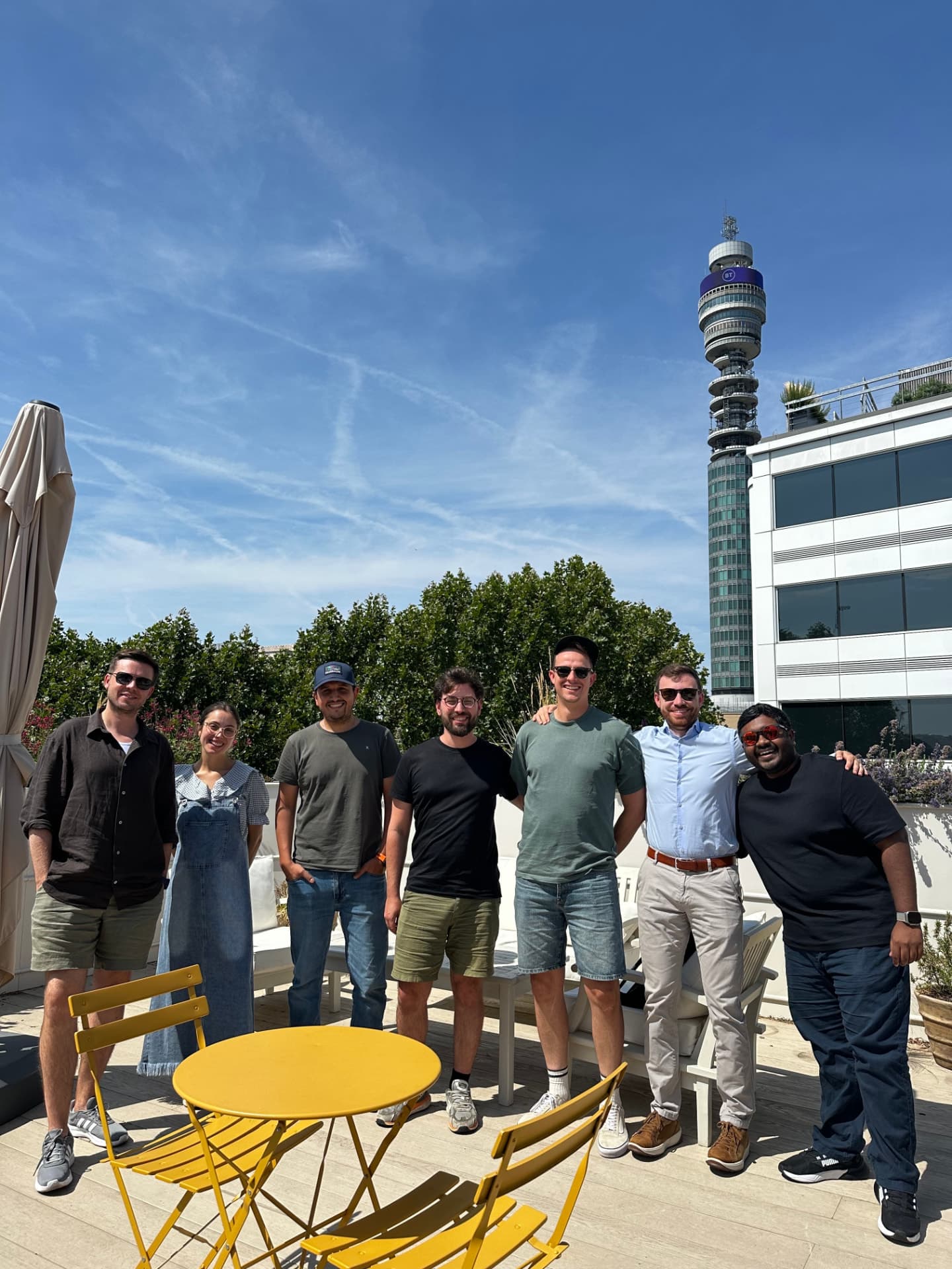 Seven of the team on a London rooftop terrace, the BT Tower behind them and yellow café chairs in the foreground.