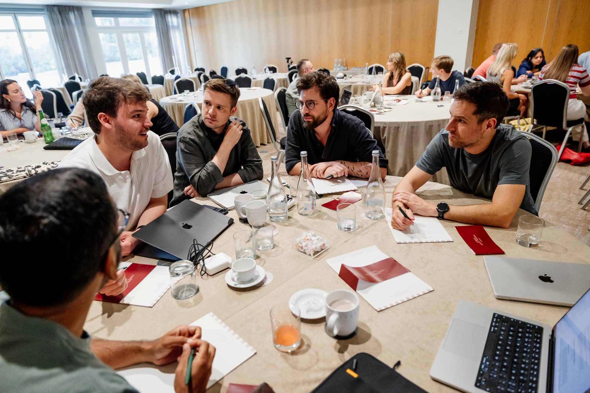 Engineers around a large round table mid-discussion, laptops, notebooks and coffee cups between them.
