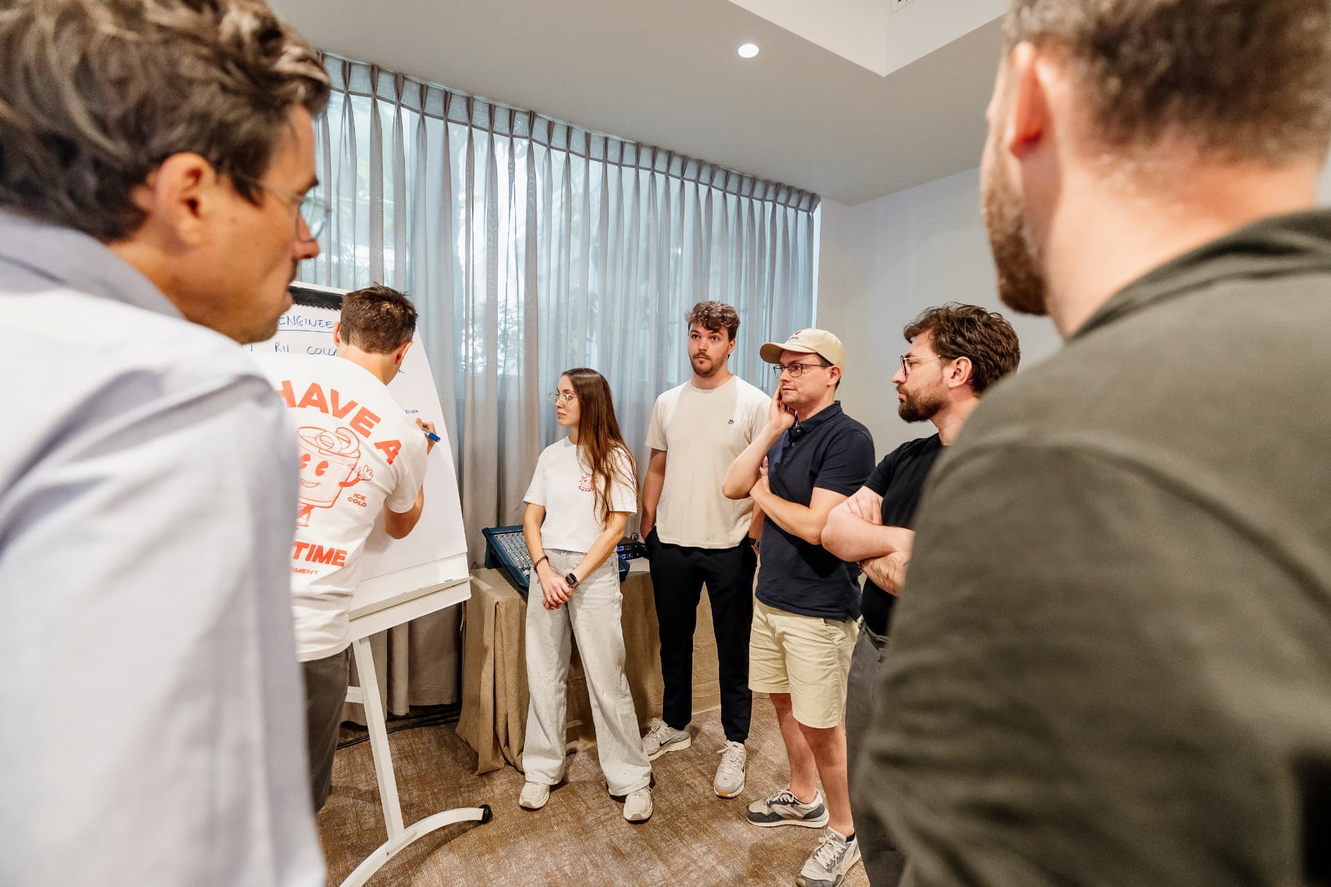 The team gathered in a bright room listening to someone present, two colleagues framing the shot in the foreground.