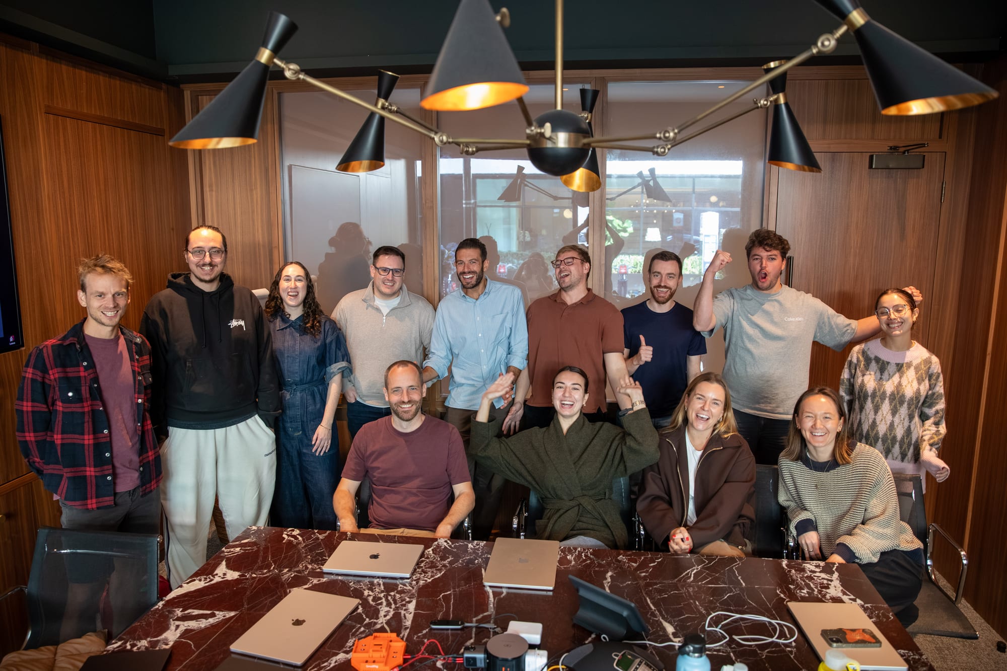 Fourteen researchers and engineers posing around a marble table in a wood-panelled meeting room in London, under a large black and brass chandelier. Several are laughing, waving or cheering with raised arms.